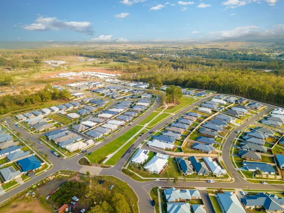 Aerial view of Huntlee Estate, a residential and industrial development with organized rows of modern homes and commercial buildings with blue roofs, connected by curved streets and green spaces, surrounded by forested hills and farmland under a partly cloudy sky.