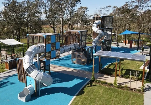 Huntlee playground featuring modern play equipment including a large spiral slide tower, tunnel slides, climbing structures, and a ping pong table on blue safety surfacing, surrounded by eucalyptus trees and shade structures on a sunny day.