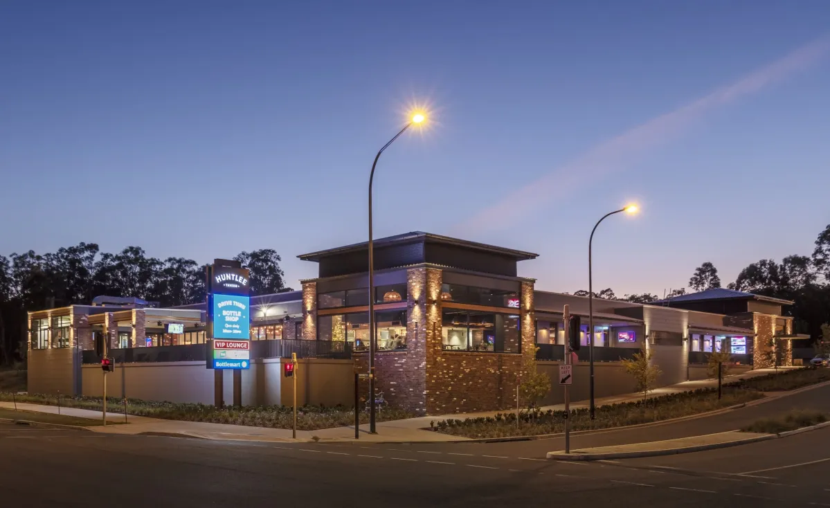 Huntlee Tavern exterior at dusk, a modern single-story building with brick and stone facade, illuminated by warm accent lighting and street lamps. The contemporary structure features large windows, a pitched roof, and signage displaying 