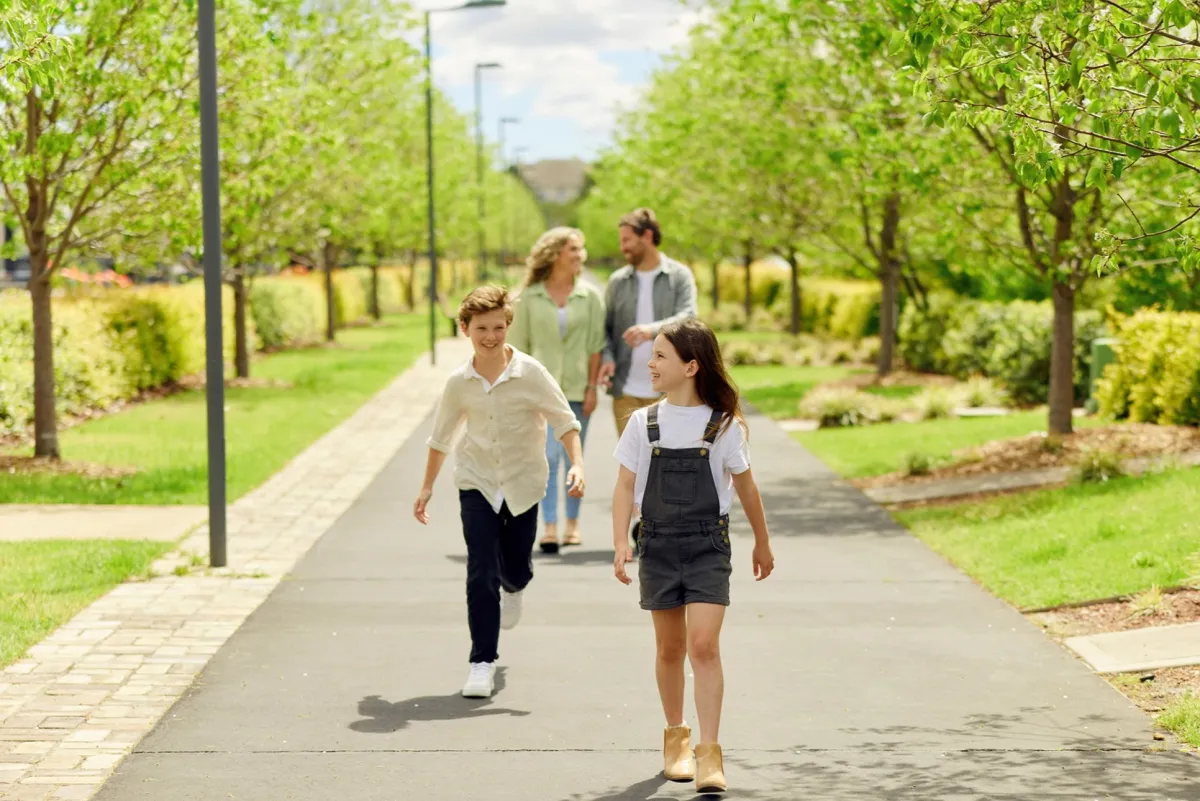 A family of four walks along a tree-lined paved pathway at Catherine Park estate in NSW. Two children run ahead on the path while two adults follow behind. The pathway is flanked by rows of young green trees on both sides, with manicured lawns and landscaping visible throughout the residential development. A modern house is visible in the distance under a partly cloudy sky.