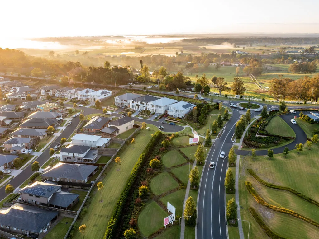 Aerial view of Red Bank residential development at sunset, showing modern white houses and established homes along curved streets, with green landscaping, a winding road, and rural farmland and waterways visible in the distance.