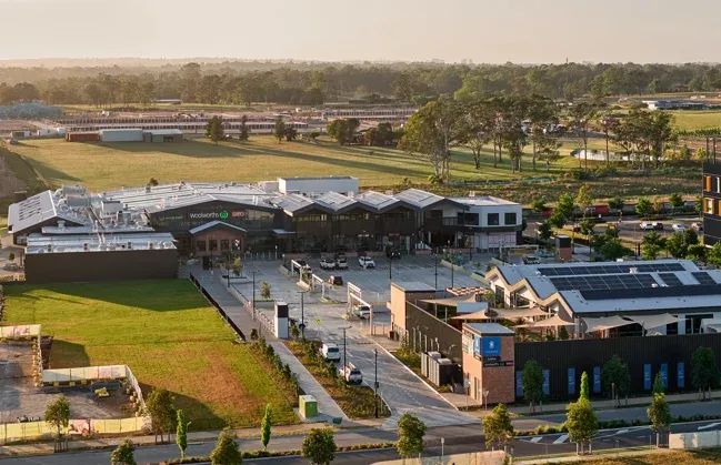 Aerial view of Stockland Gables town centre showing modern commercial buildings with metal and brick facades, green landscaped grounds, parking areas, and surrounding residential neighborhoods with trees and open spaces.