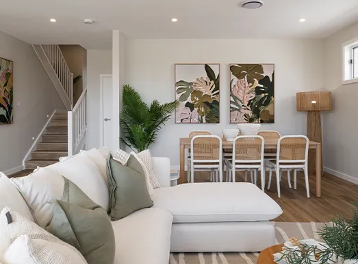 Modern living room and dining area with cream-colored sofa with green and white pillows, white ottoman, wooden dining table with wicker-backed chairs, botanical wall art, potted plants, wooden floor, white staircase visible in background, and recessed ceiling lights.