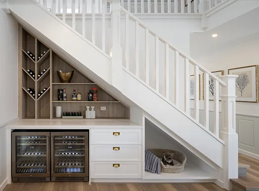Modern staircase with built-in storage featuring wine coolers, white cabinetry, decorative shelving with wine glasses and decor items, and open storage niche with woven basket underneath the stairs.