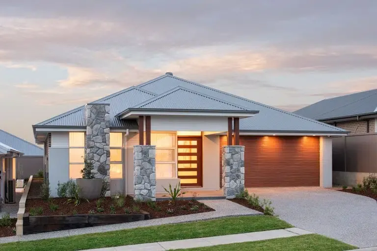 Modern single-story display home with white exterior walls, metal roof, stone pillars framing the entrance, illuminated wooden front door, garage with brown roller door, landscaped front garden with lawn and shrubs, photographed at dusk in Huntlee.