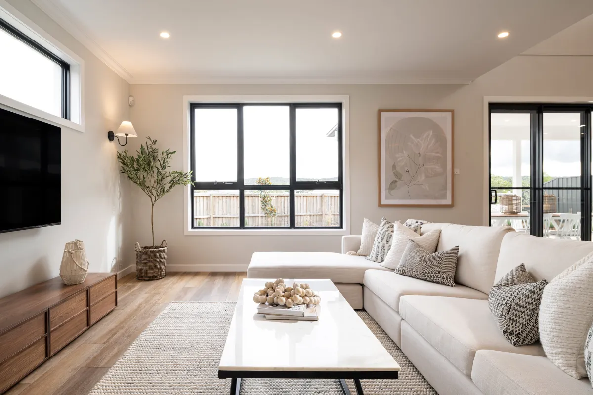 Modern living room with cream-colored L-shaped sofa, white coffee table with decorative bowl, and patterned throw pillows. Wood flooring and neutral area rug. Large black-framed windows and glass doors overlook balcony. Wall-mounted TV on left, wooden media console below. Botanical artwork above sofa. Potted plant, wall sconce, and recessed ceiling lights complete the contemporary design.