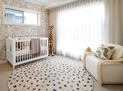 A modern nursery room with a white wooden crib against patterned wallpaper, a beige upholstered chair with decorative pillows on the right, a patterned area rug with geometric designs, sheer curtains covering large windows allowing natural light, and decorative giraffe figurine on the floor.