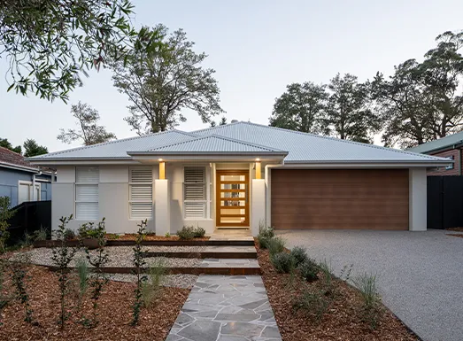 Modern single-storey home with white exterior walls and grey metal roof, featuring a wooden front door with sidelights, brick driveway, landscaped front garden with native plants, and attached garage on the right side, set among tall trees on Central Coast.