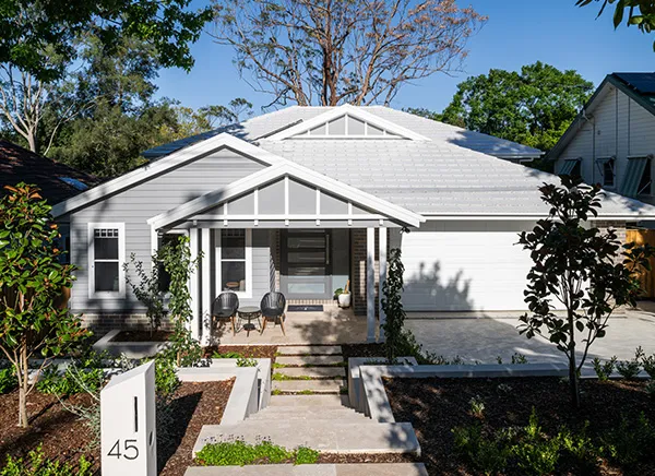 Modern white single-story house with gabled roof, front porch with columns, garage, concrete driveway, and landscaped garden with young plants and shrubs under blue sky.