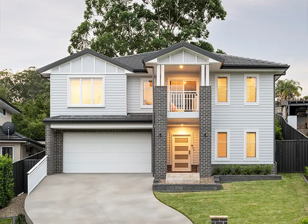 Modern two-story residential home in Sydney featuring white weatherboard cladding with charcoal brick accents, double garage, front portico with white railings, and illuminated windows at dusk. Mature trees visible in background with manicured front lawn.
