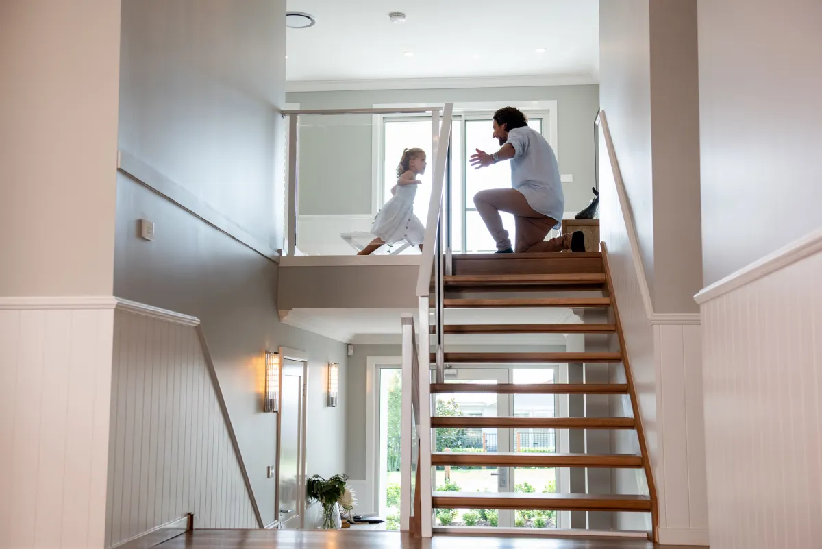 Adult and child playing on wooden staircase in a modern two-story home with white walls, glass railings, and natural light from large windows.