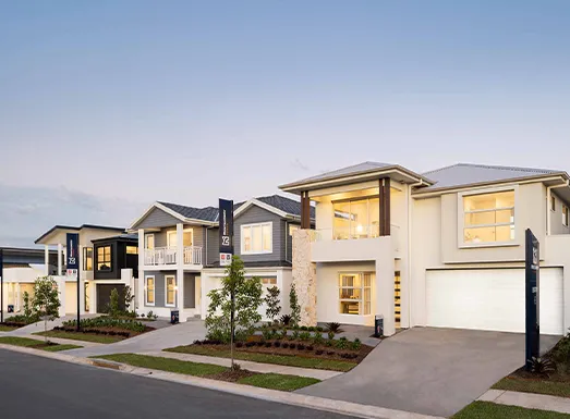 Modern residential street showing two contemporary two-story houses with cream and dark gray facades, large illuminated windows, manicured front landscaping, driveways, and a clear evening sky.