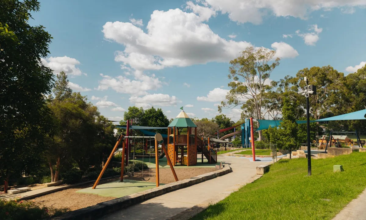 Millfield Rise Estate playground featuring a large multi-level play structure with green roof tower, orange and red climbing elements, and slides, surrounded by landscaped grounds with grass, trees, and blue shade sails on a sunny day.