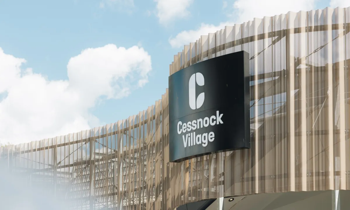 Exterior of Cessnock Village shopping center with modern corrugated metal and timber facade under blue sky with white clouds. Black signage with white logo and text reading 
