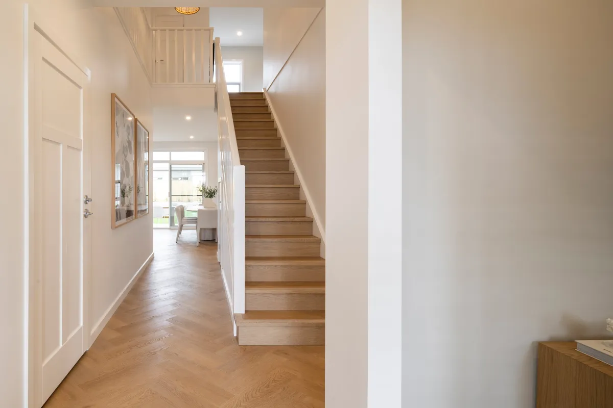 Modern home entryway with wooden staircase, white paneled doors on left wall, wooden flooring, and view through to living areas with natural light from windows.