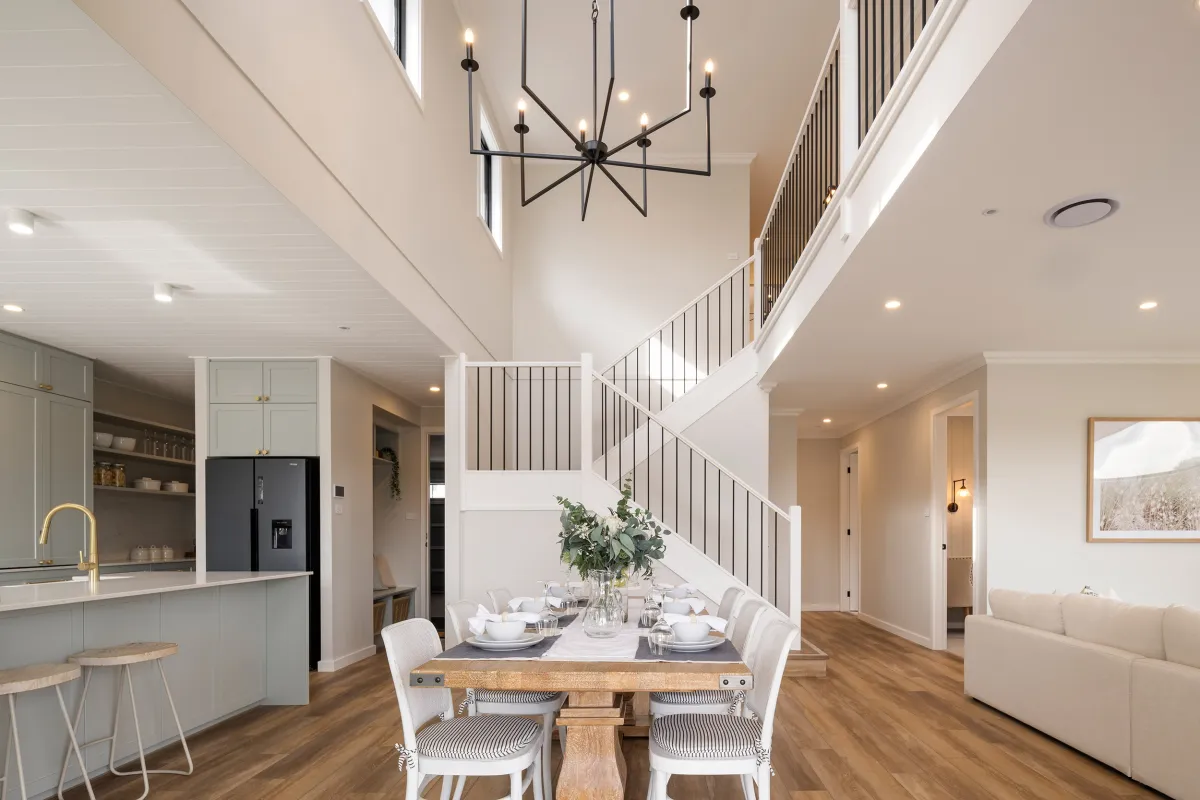 Modern open-plan dining and kitchen area in a display home featuring a wooden dining table set for six with white upholstered chairs, a contemporary black geometric pendant light fixture overhead, light gray cabinetry with brass hardware, black refrigerator, and a curved white staircase with metal railings visible in the background, all with warm wood flooring and neutral décor.
