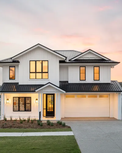 Montgomery Homes Waverley display home exterior at dusk. Modern two-story house with white board and batten siding, black metal roof, and large illuminated windows. Black front door with sidelights, two-car garage, manicured lawn, and landscaping. Soft pink and golden sunset sky in background.
