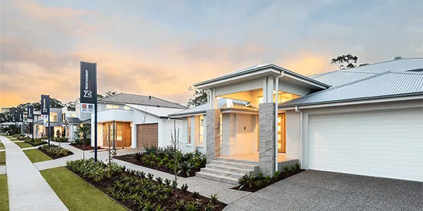 Row of modern display homes at dusk with illuminated interiors, featuring contemporary architecture with white facades, metal roofing, and manicured front landscaping along a residential street in Newcastle.