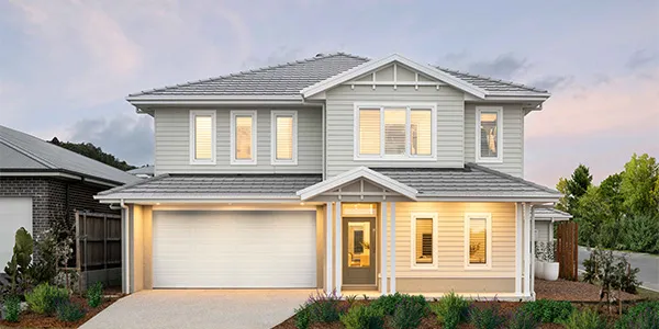 Modern two-story residential home with light gray and white exterior, featuring a double garage, cream-colored front door, multiple windows with warm lighting, pitched roof with gray tiles, concrete driveway, and manicured front landscaping at dusk.