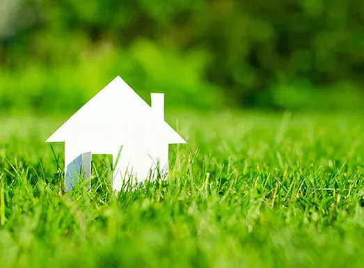 White paper house cutout placed on green grass with blurred green foliage background, representing property search or finding a home location.