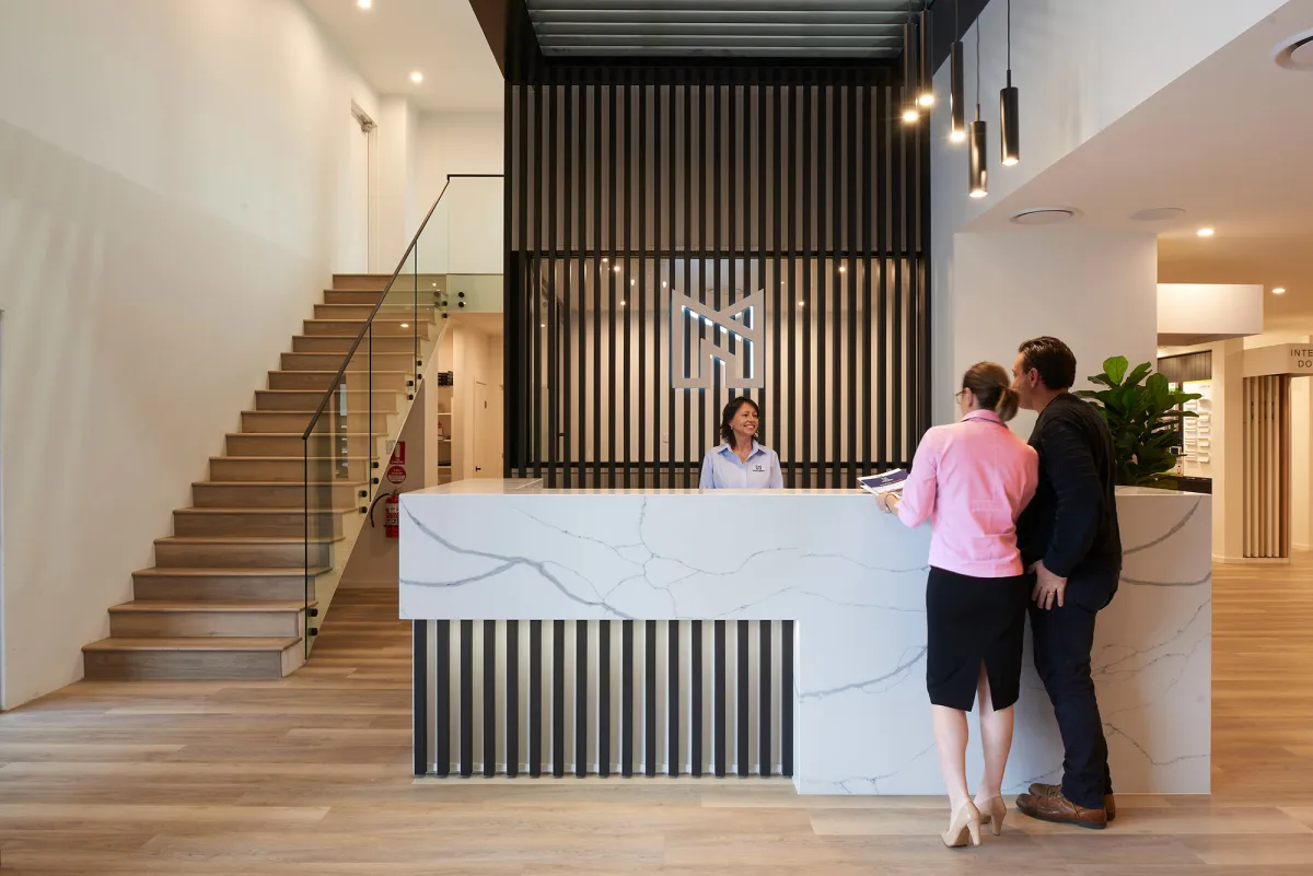 Modern office lobby with a white marble reception desk featuring vertical black slat paneling behind it. A receptionist in a blue shirt greets two visitors—a woman in pink and a man in black. Wooden stairs with glass railing are visible on the left, and the space features warm lighting, wood flooring, and contemporary design elements including a potted plant and pendant lights.