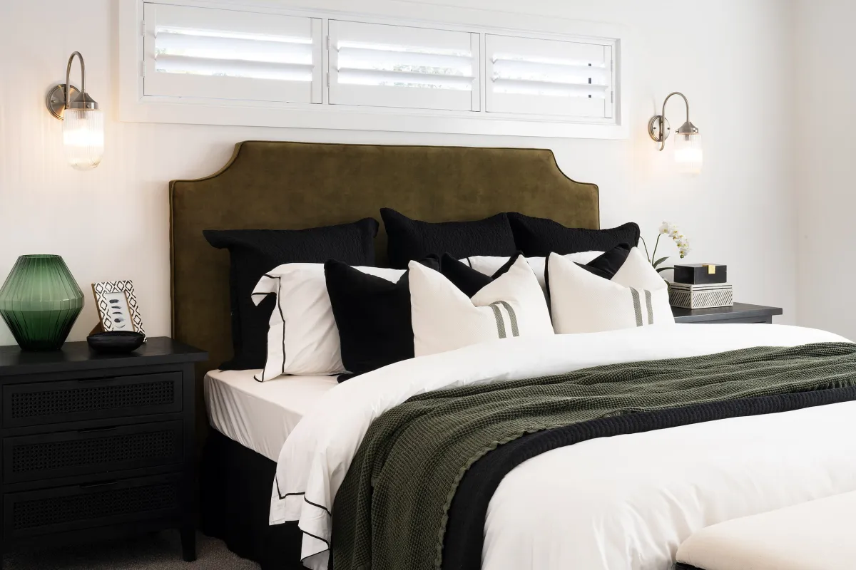 Modern bedroom with olive green upholstered headboard, white bedding with black and cream pillows, olive and black throw blanket, black nightstand with green glass lamp and decorative items, brass wall sconces on either side of the bed, and transom windows above.