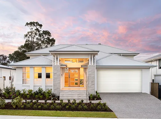 Modern white single-story house with metal roof, featuring a central entrance with warm-lit wooden doors and sidelights, attached garage on the right, manicured landscaping with low shrubs, concrete driveway, and tree in background against a pink and purple sunset sky.