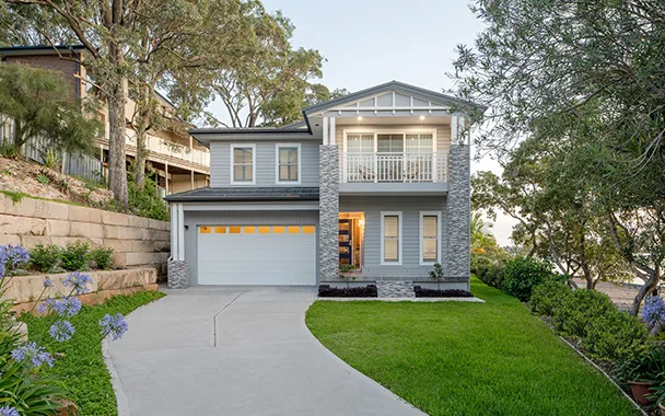 Modern two-story residential home with light gray and blue exterior, featuring a double garage with yellow-lit door, manicured lawn, curved driveway, retaining wall, and mature trees surrounding the property on the Central Coast.