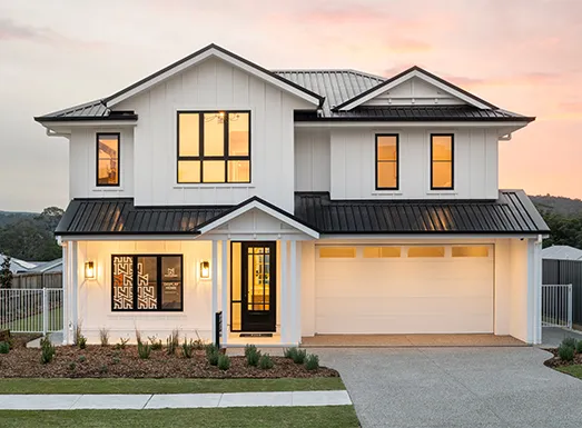 Modern two-story white house with black metal roof and trim, illuminated windows, black front door, and garage, set against a pink sunset sky with landscaped front yard and white fence.