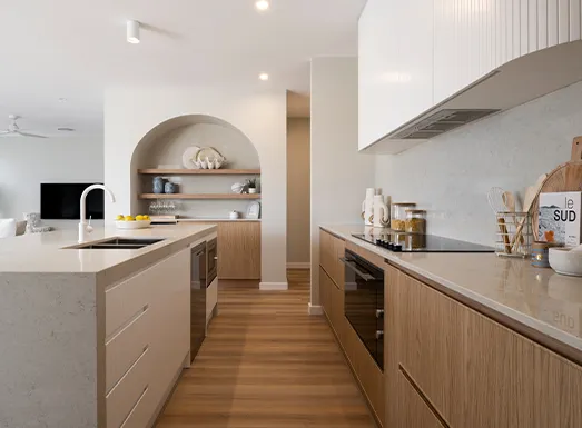 Modern kitchen with light wood cabinetry, white countertops, and stainless steel appliances. Features an arched niche with shelving on the back wall, island bench with seating, pendant lighting, and wooden flooring throughout.