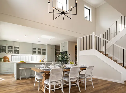Modern open-plan kitchen and dining area with white cabinetry, wooden dining table with upholstered chairs, stainless steel appliances, and a staircase with black metal railings visible in the background.