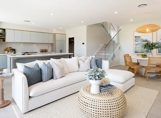 Modern open-plan living space in Wollongong featuring a white sectional sofa with decorative pillows, round woven ottoman coffee table, light wood dining chairs, contemporary kitchen with white cabinetry, metal staircase, and neutral color palette with natural lighting.