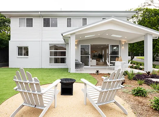 Modern white residential home with covered patio and backyard entertaining space, featuring two white Adirondack chairs facing a fire pit on sandy ground, manicured lawn, and landscaping in Wollongong.