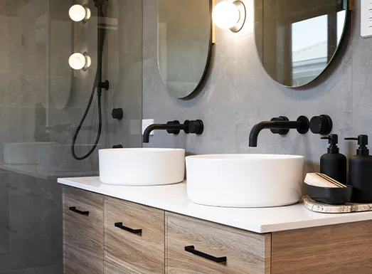 Modern bathroom vanity with dual white cylindrical sinks, black faucets, and rounded mirrors with sconces above. Wooden cabinetry with black handles sits beneath a white countertop. Glass shower enclosure visible on the left with rainfall showerhead and black fixtures.