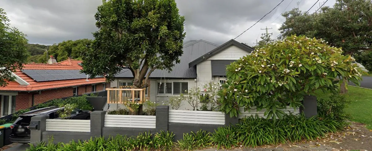 Merewether residential property before renovation showing a weathered cream and dark gray house with metal roof, fronted by a modern gray and white fence, mature trees, and overgrown garden vegetation under overcast sky.