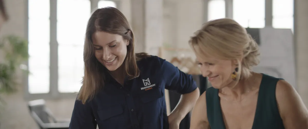 Two women smiling while looking at a laptop in a bright, modern office space with large windows and plants in the background. The woman on the left wears a dark blue branded polo shirt, while the woman on the right wears a dark green sleeveless top.