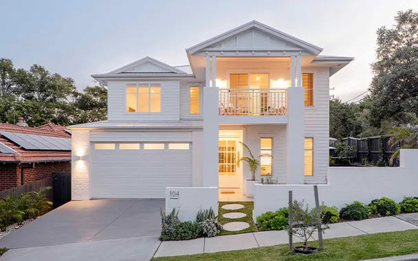 Modern two-story white house with contemporary design, featuring a double garage, large windows with warm interior lighting, front balcony, landscaped garden with stepping stones, and mature trees in the background at dusk.