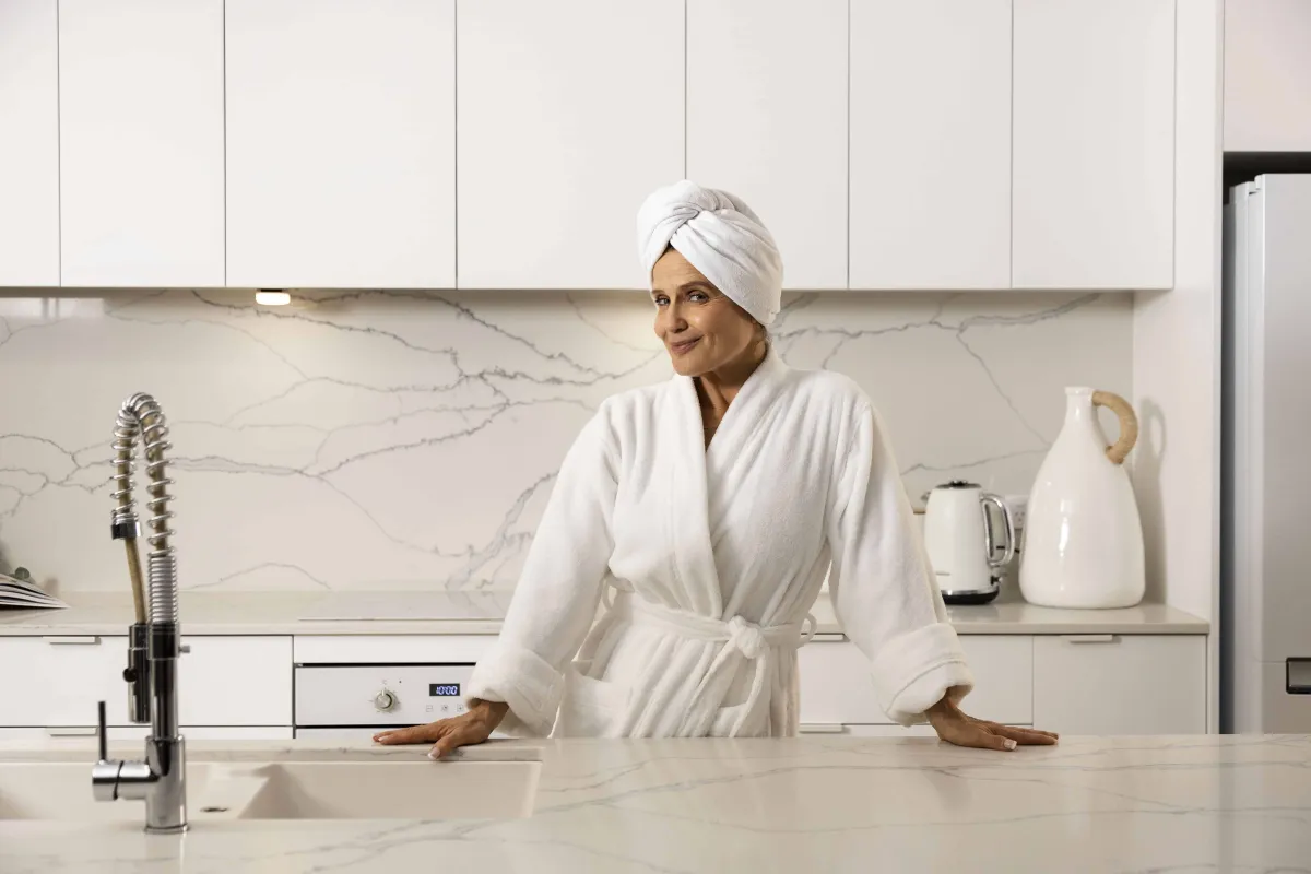 Shaynna Blaze in white bathrobe and towel wrap standing at modern kitchen counter with marble backsplash, white cabinets, stainless steel faucet, and white appliances.