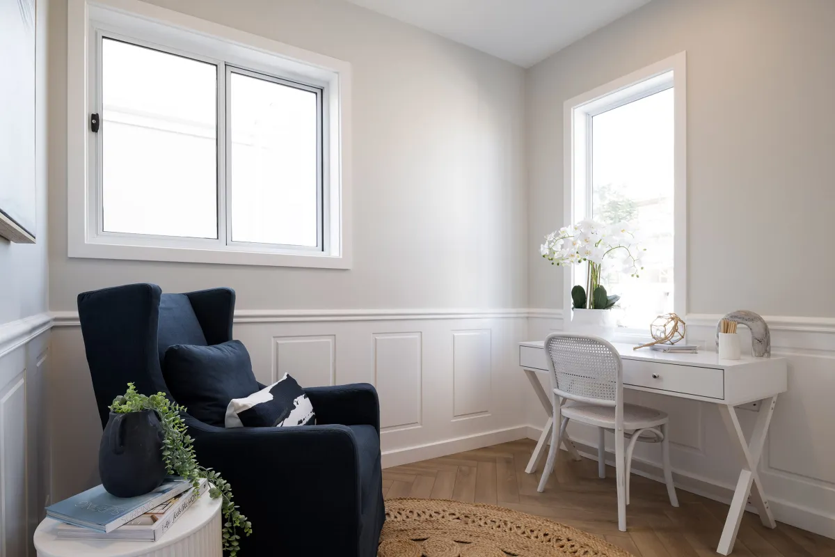 Modern bedroom interior with a dark navy armchair near a window on the left, a white desk with chair by a bright window on the right, light beige walls with white paneling, wooden flooring, and decorative elements including white orchids and potted plants.