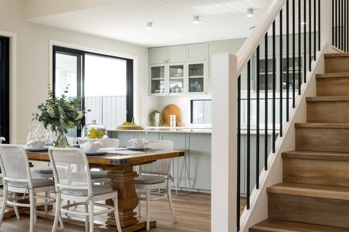 Modern farmhouse dining area with wooden table and white mesh chairs, adjacent to light gray cabinetry with glass doors and brass hardware. A vase of fresh flowers and place settings sit on the table. Black and white staircase with wooden treads visible on the right. Large black-framed glass doors open to a bright balcony. Recessed lighting in white ceiling and potted plants complete the bright, contemporary farmhouse aesthetic.
