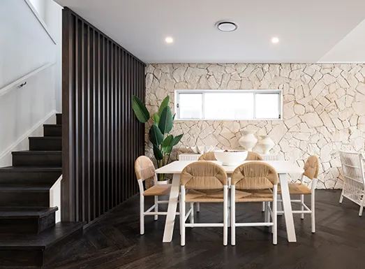 Modern dining area with white table and natural wood chairs, beige stone accent wall, dark vertical curtains, black staircase on left, potted plant, and recessed ceiling lights in contemporary home interior.