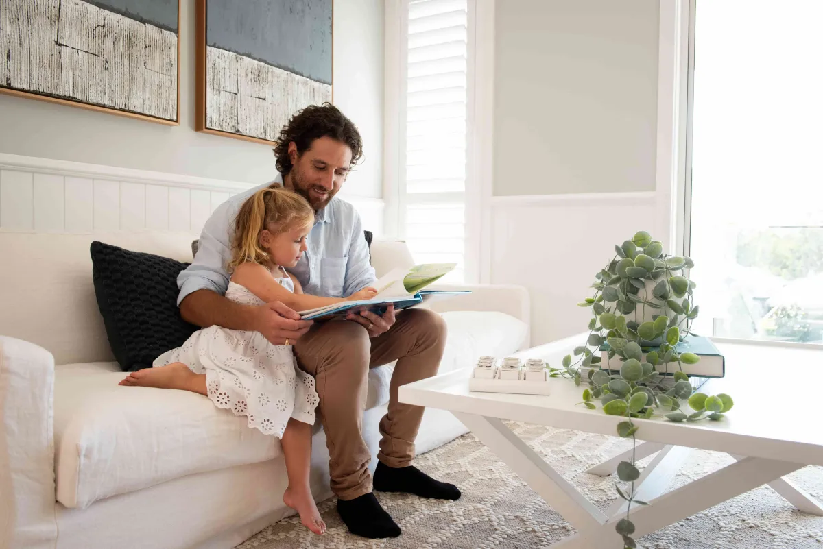 A man and young child sit together on a cream-colored sofa reading a picture book in a bright, modern living room. Sunlight streams through large windows behind them, illuminating white shiplap wall paneling and abstract artwork. A potted succulent plant and decorative books sit on a side table nearby.
