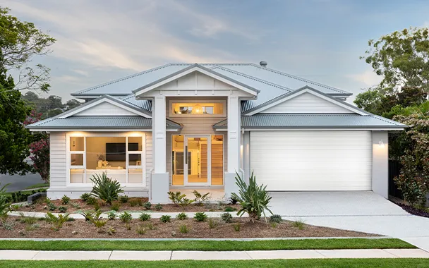 Modern white single-story house with blue metal roof, large garage door, and illuminated front entrance with warm lighting. Landscaped front yard with native plants and green lawn. Contemporary architectural style typical of Australian knock-down rebuild homes.
