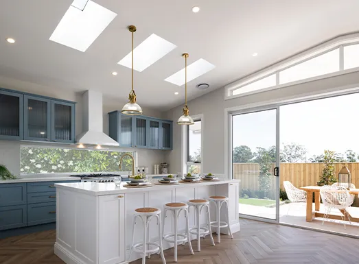 Modern kitchen with white island and bar seating, blue cabinetry on left, skylights overhead, pendant lighting, stainless steel appliances, and large glass doors opening to backyard with wooden deck at Oran Park.
