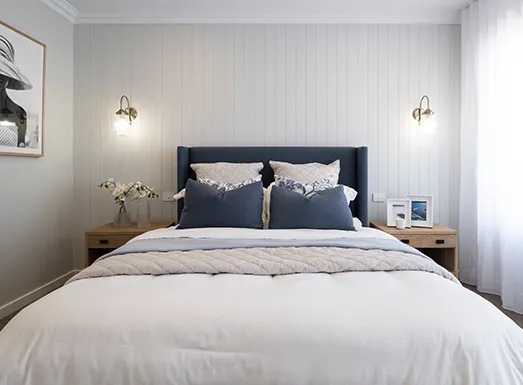 Modern bedroom with a navy upholstered headboard, white bedding with decorative pillows, wooden nightstands with brass sconce lighting on either side, white vertical shiplap walls, and soft natural light from a window with white curtains.