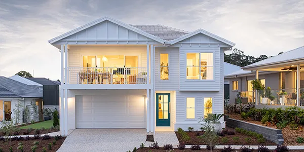 Modern two-story display home with light blue exterior cladding, white garage door, and gabled roof. Front facade features large windows with warm interior lighting, a covered upper balcony, and landscaped front garden with mulch beds. Neighboring homes visible on either side. Taken at dusk with cloudy sky.