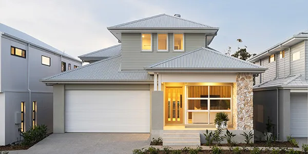 Front facade of Savannah 262 display home in Oran Park, featuring a modern two-story house with grey rendered walls, metal roof, white garage door, timber-framed entry with stone accent column, and illuminated windows at dusk.