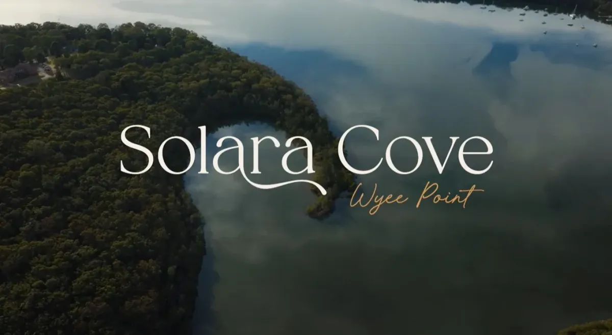 Aerial view of Solara Cove at Wyee Point, showing a forested peninsula jutting into calm blue water surrounded by dense green trees under overcast skies.