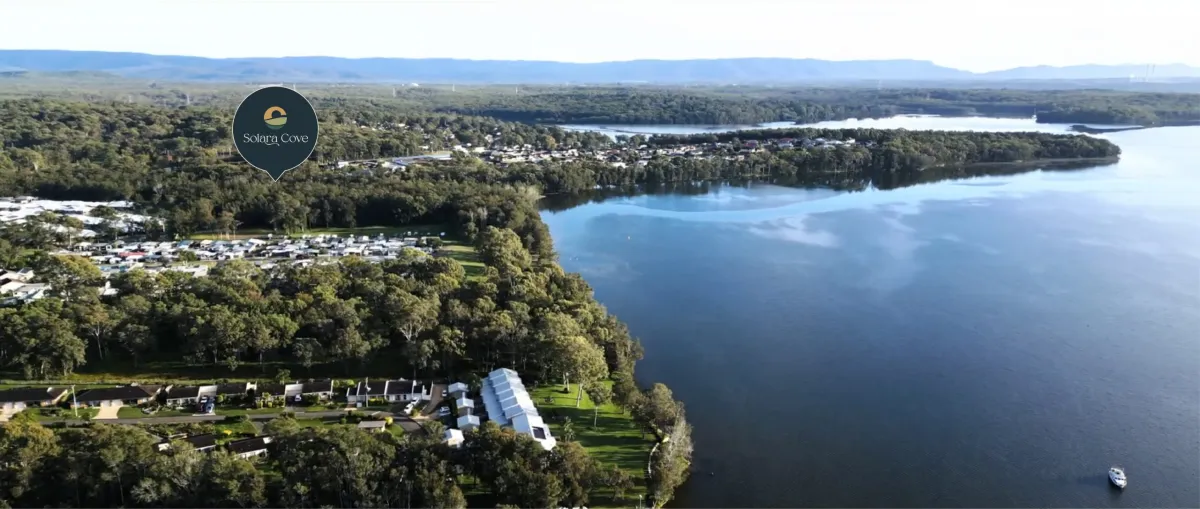 Aerial view of Solara Cove location marked with a pin on a forested peninsula overlooking a large blue lake, with residential buildings and a community center visible among green trees, surrounded by water and distant hills.