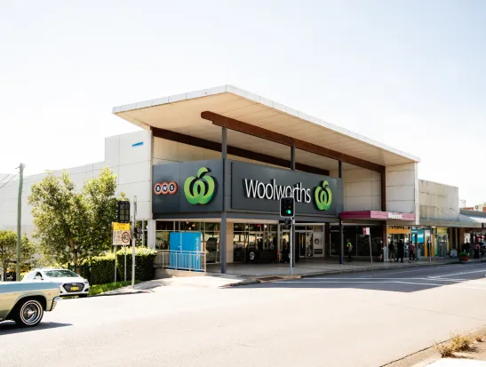 Solara Cove shopping center exterior featuring a modern Woolworths supermarket with green logo, large storefront windows, and wooden canopy overhang. Additional retail shops visible on either side of the main entrance, with parking area and landscaping in foreground.